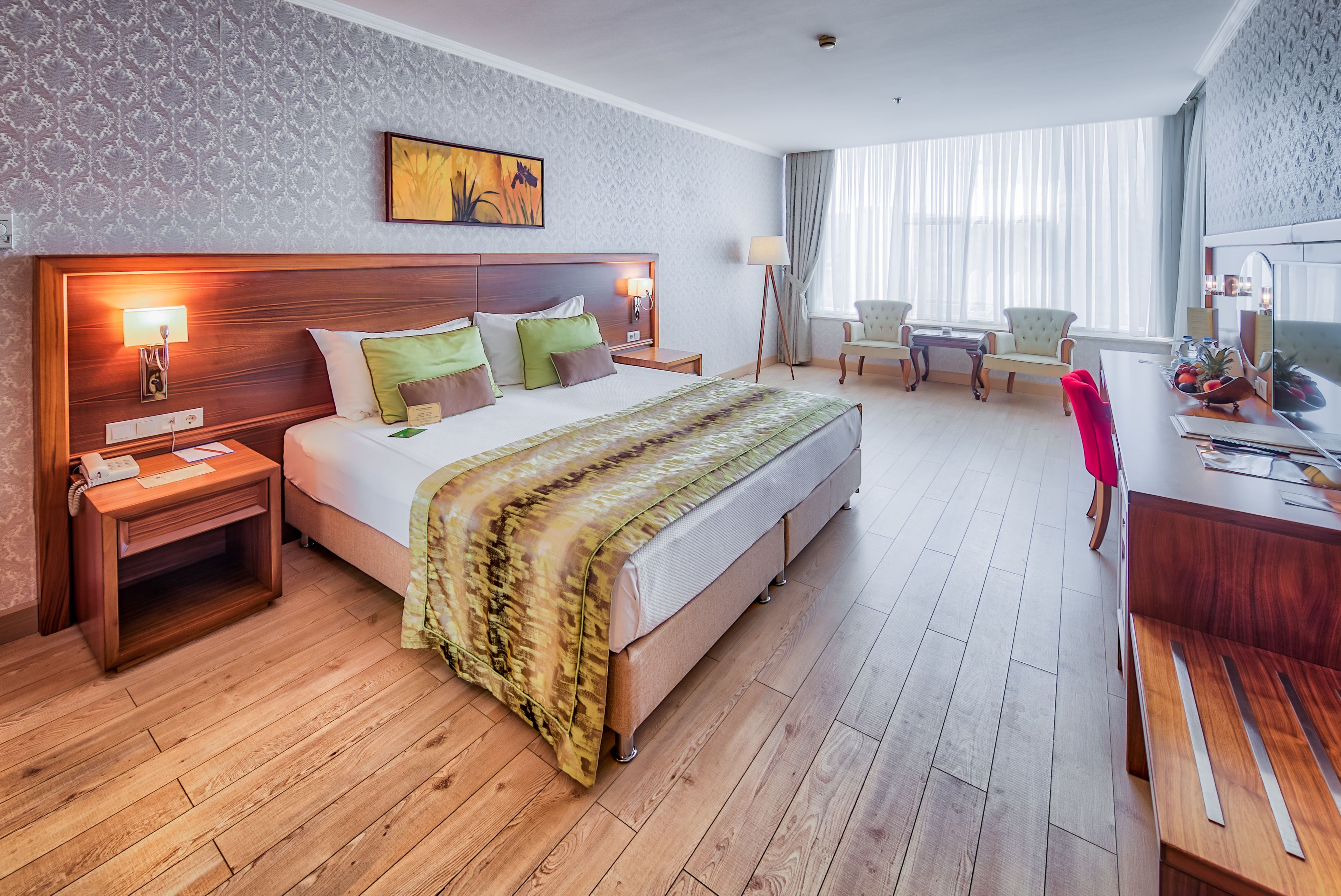 A hotel room with a large bed, wooden headboard, bedside tables, a desk with a chair, and a large window with curtains.