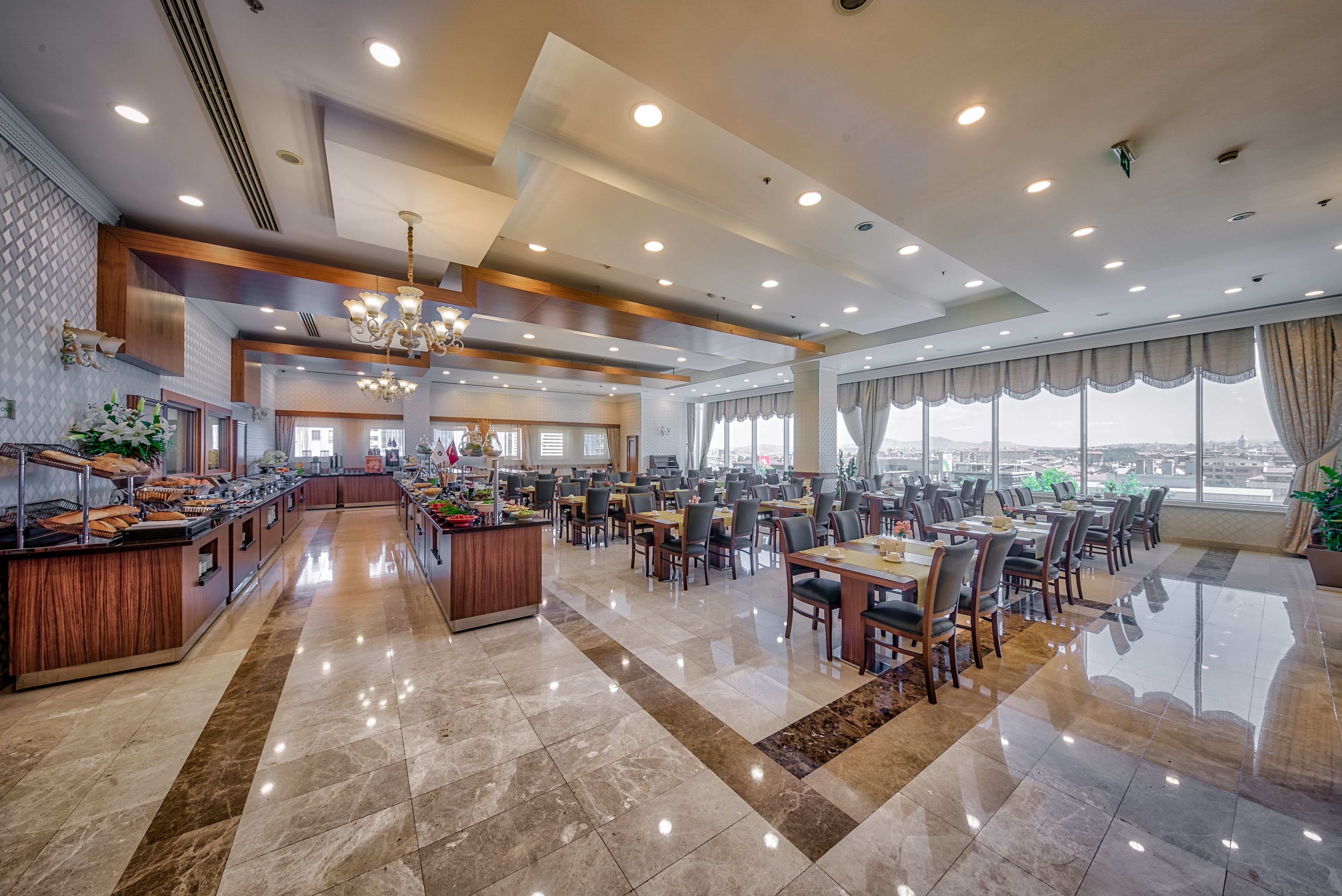 Amplio comedor de hotel con numerosas mesas y sillas, una estación de buffet central y grandes ventanales con vistas a la ciudad.