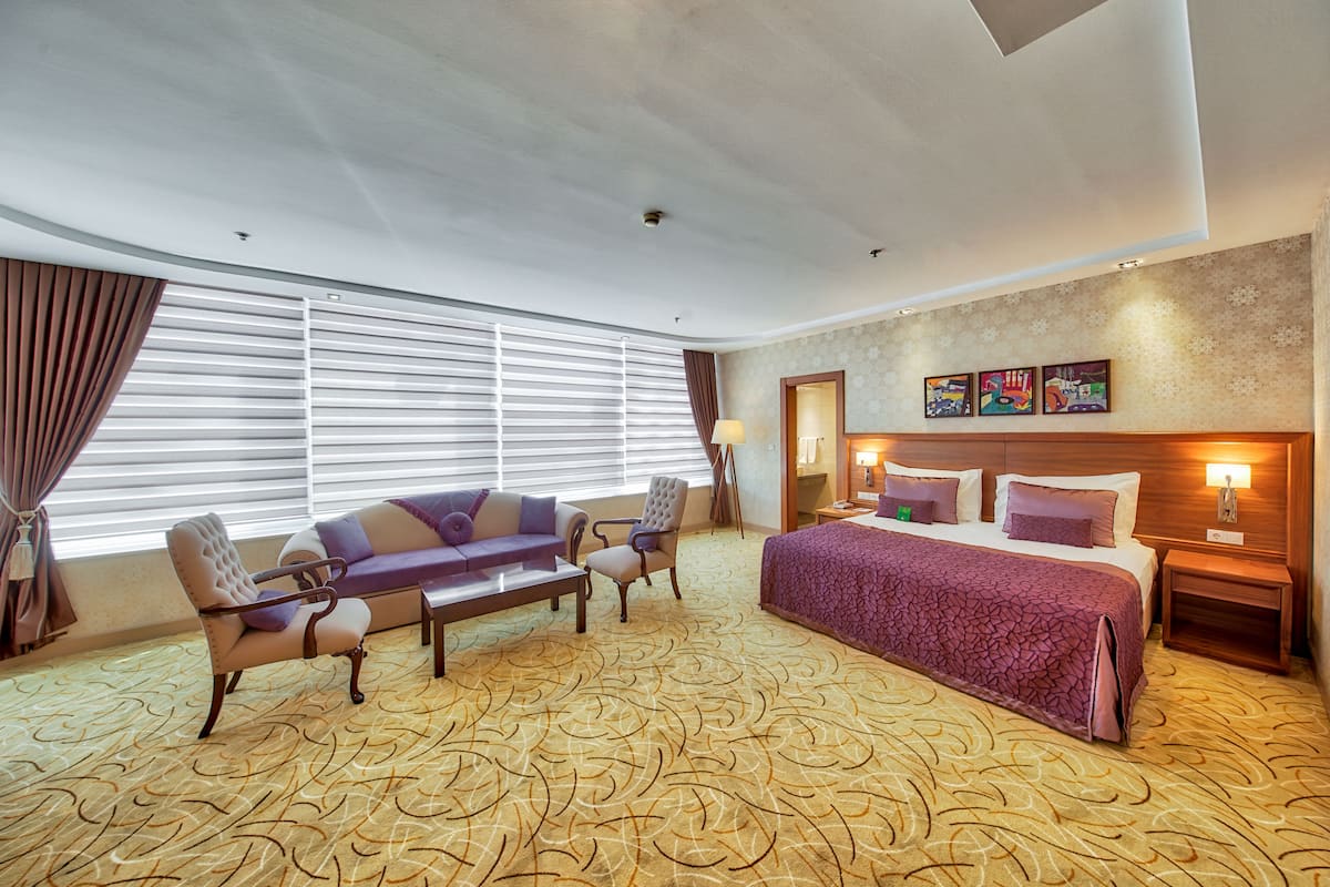 Habitación de hotel con una cama grande, dos sillones, un sofá, una mesita y un ventanal con persianas.