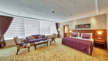 A hotel room with a large bed, two armchairs, a sofa, a small table, and a large window with blinds.