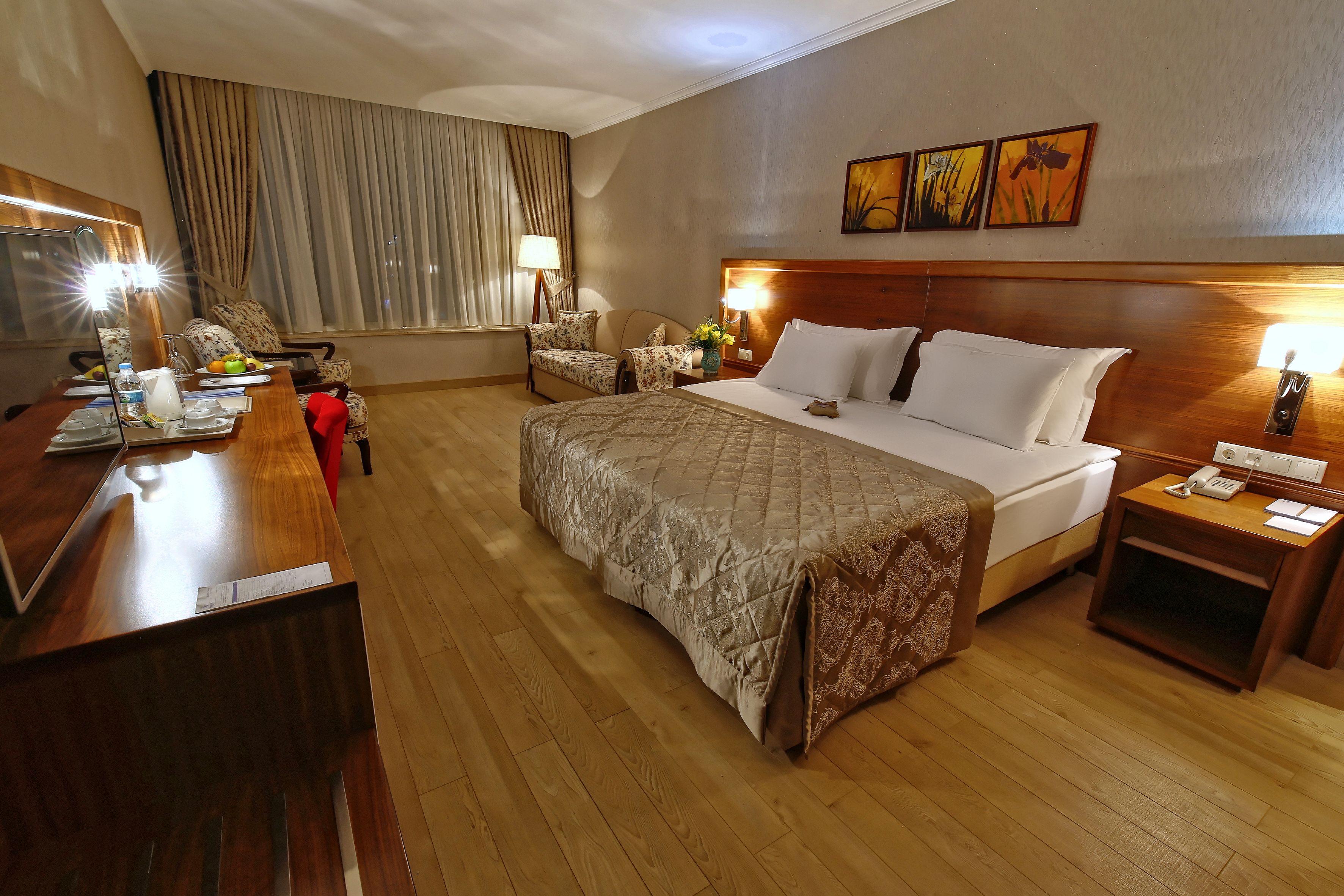 A hotel room with a large bed, a wooden desk, a chair, a bedside table with a lamp, and a window with curtains.