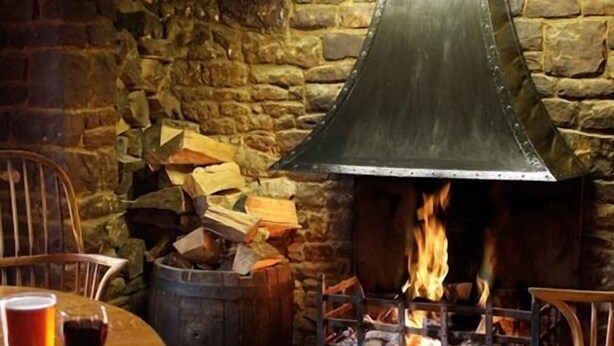 Eine Steinsäule mit Kamin aus Metall, einem Holzfässchen und einem Tisch mit einem Glas Rotwein.