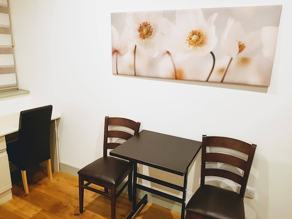 A small dining area with a table and two chairs, a white wall, and a painting of flowers. - Forest House Hotel (London)