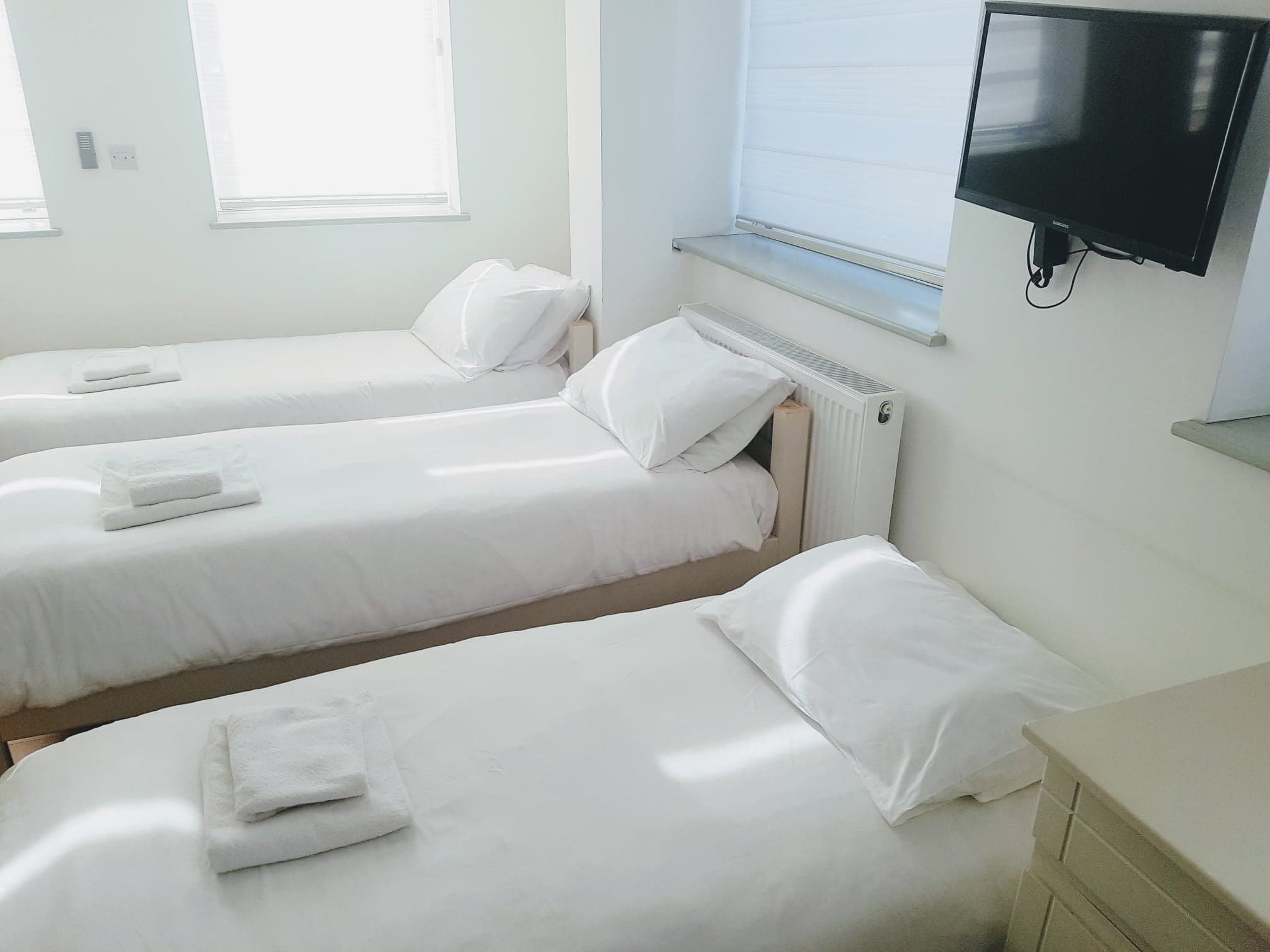 A hotel room with two single beds, white bedding, a flat-screen TV mounted on the wall, and a window with blinds.