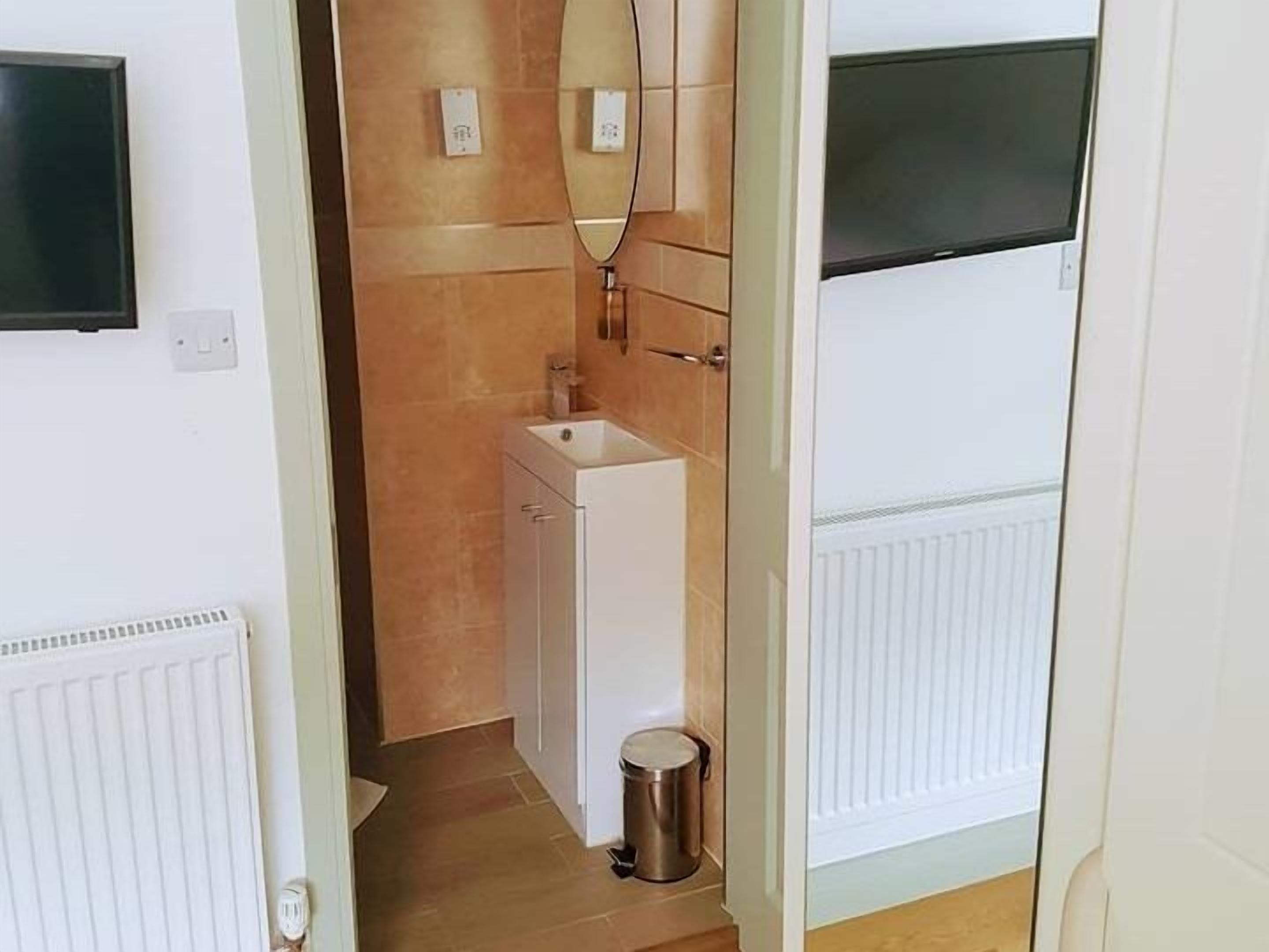 Een smalle gang met een witte radiator, een klein wastafeltje met spiegel en een prullenbak.