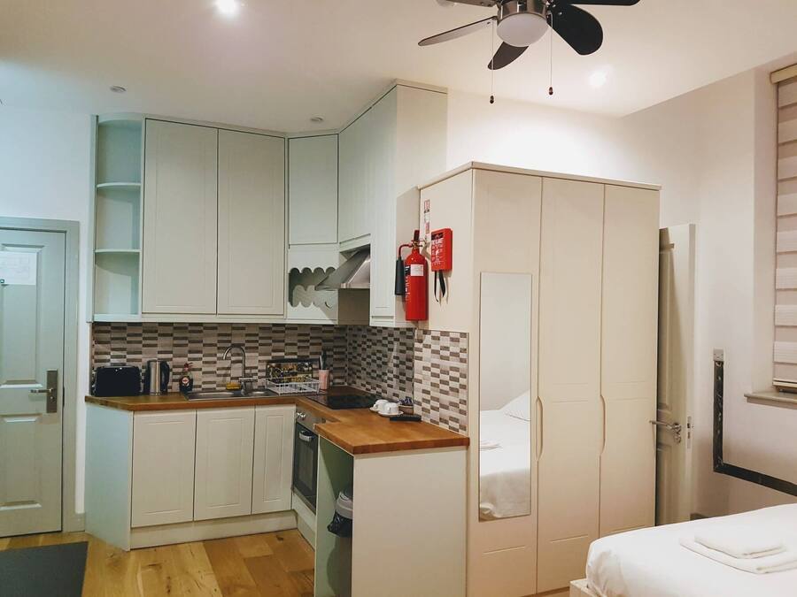 A compact kitchen with white cabinets, a microwave, and a toaster. There is a ceiling fan, a fire extinguisher, and a small window with blinds.