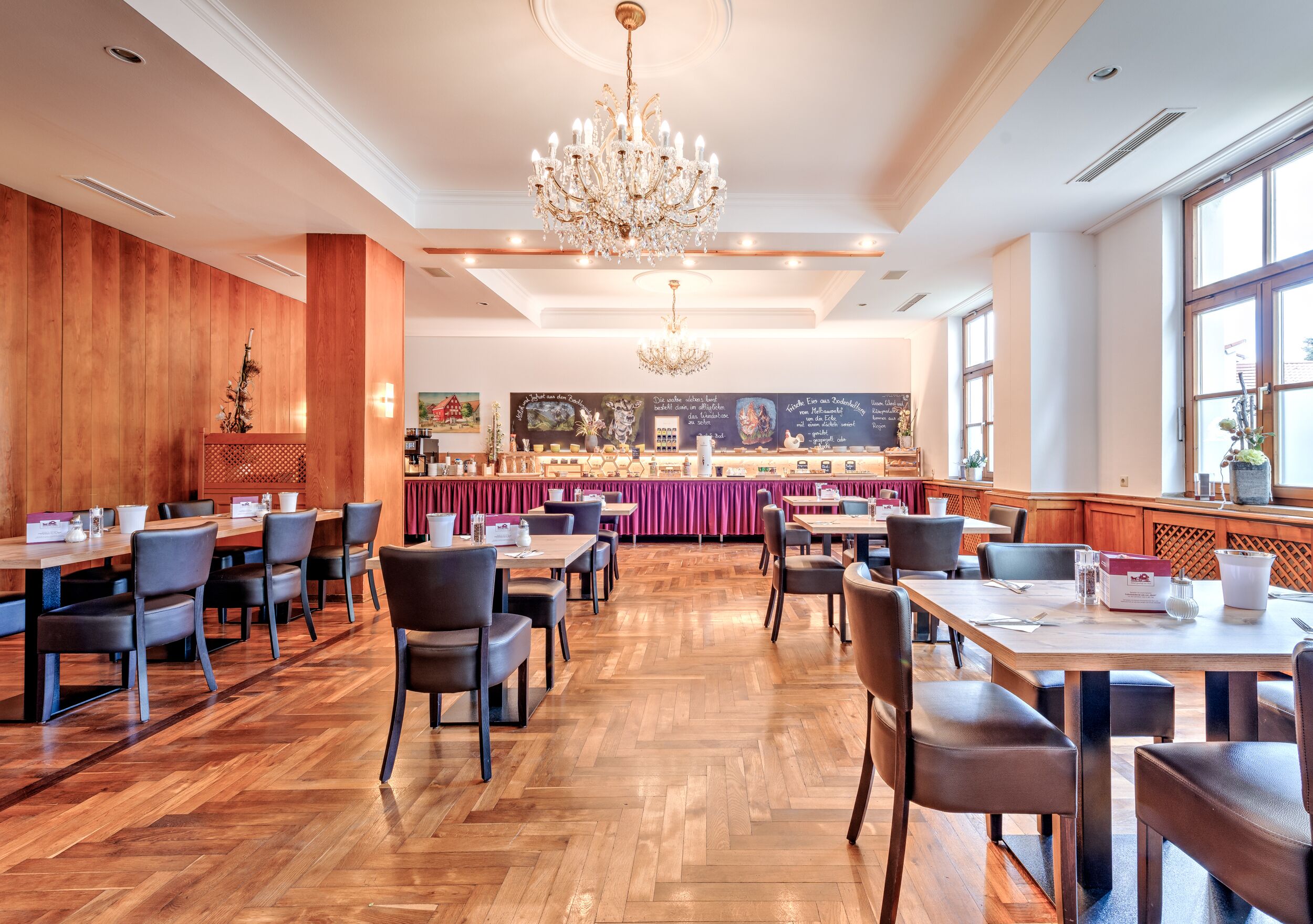 A spacious restaurant with wooden flooring, multiple tables and chairs, a central bar area, and a chandelier.