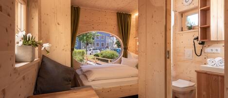 Ein Zimmer aus Holz mit einem Bett, einem kleinen Tisch, einem Fenster mit Blick auf Gebäude und einem Badezimmer mit Toilette und Waschbecken.