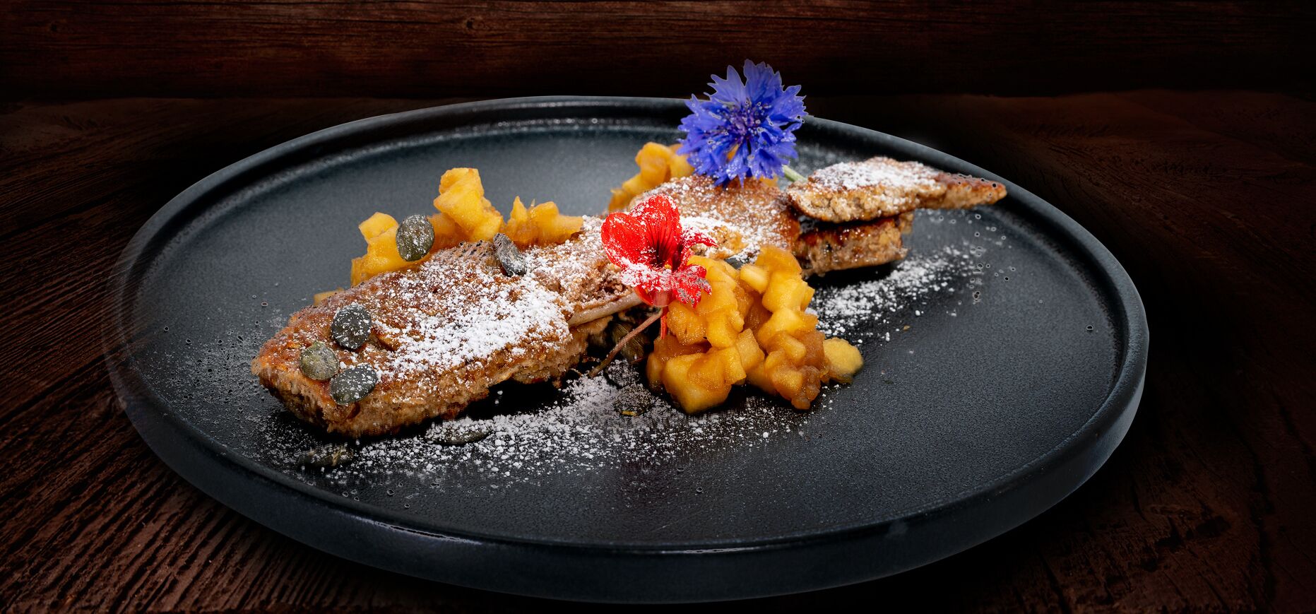A dessert plate with a pastry, fruit, and a blue flower decoration on a dark plate.