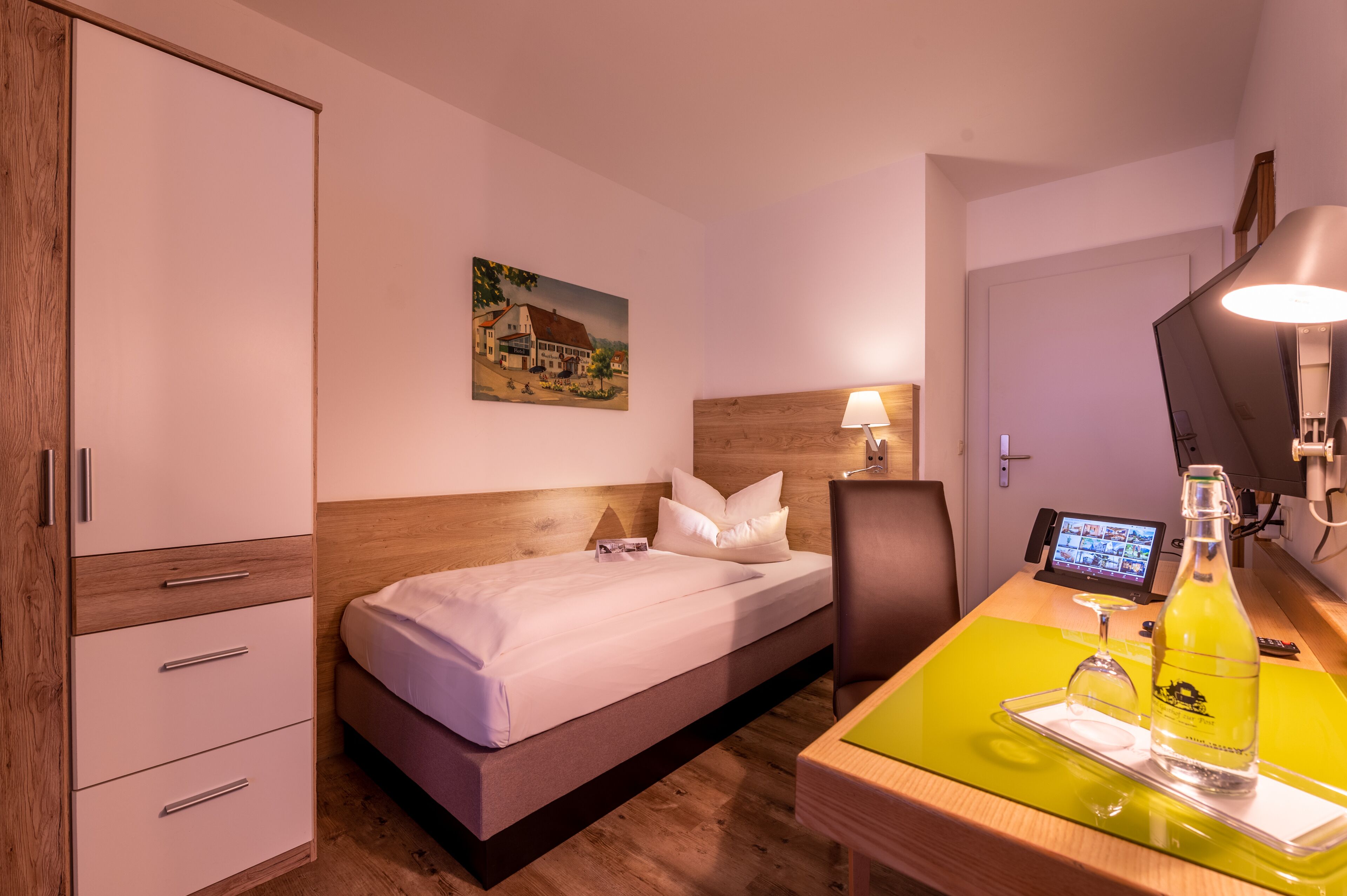 A hotel room with a bed, a desk with a lamp, a bottle, and a painting on the wall.