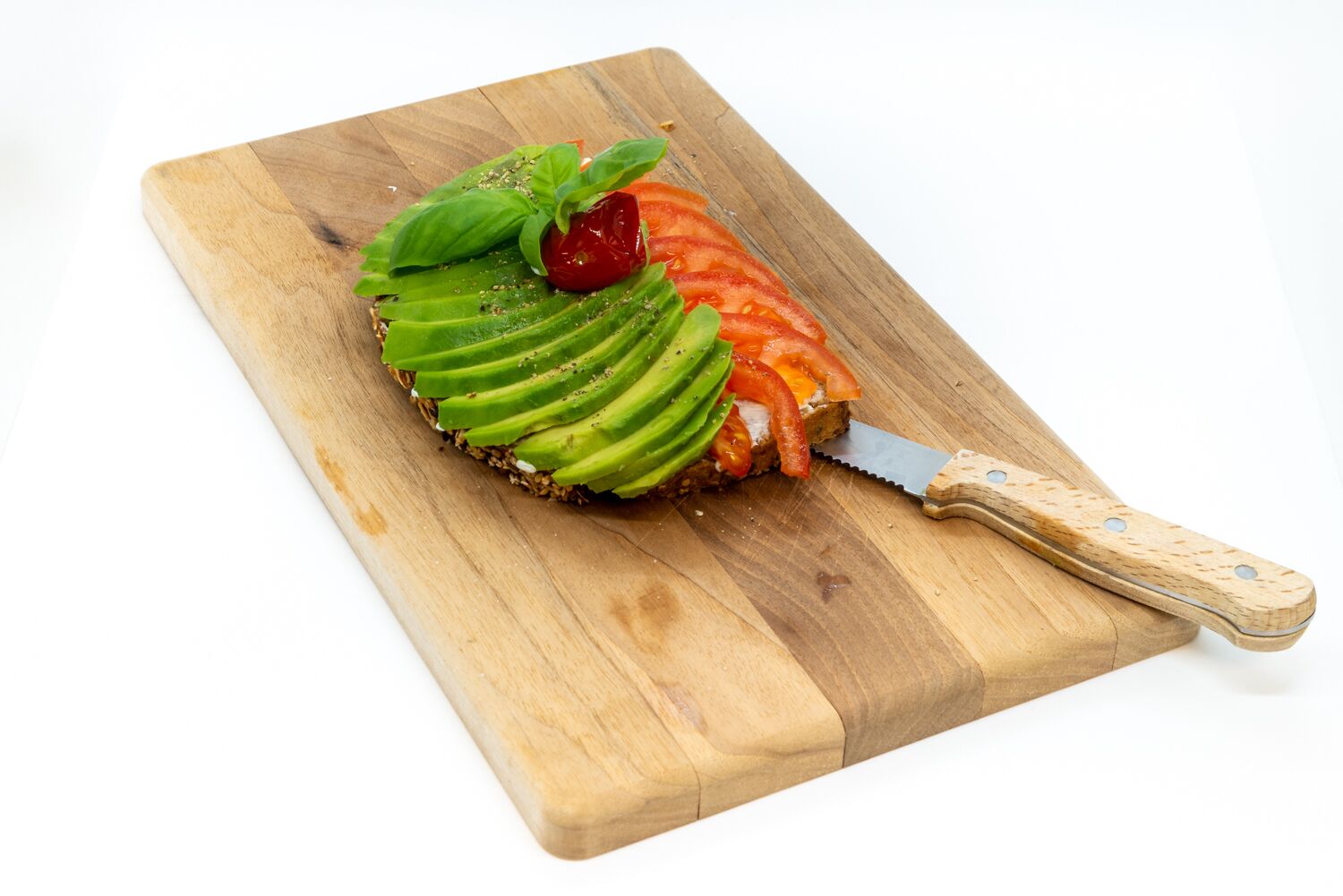 Ein Holzschnittbrett mit Lachs, belegt mit Avocado-Scheiben, Tomaten und Basilikum, sowie ein Messer mit Holzgriff.