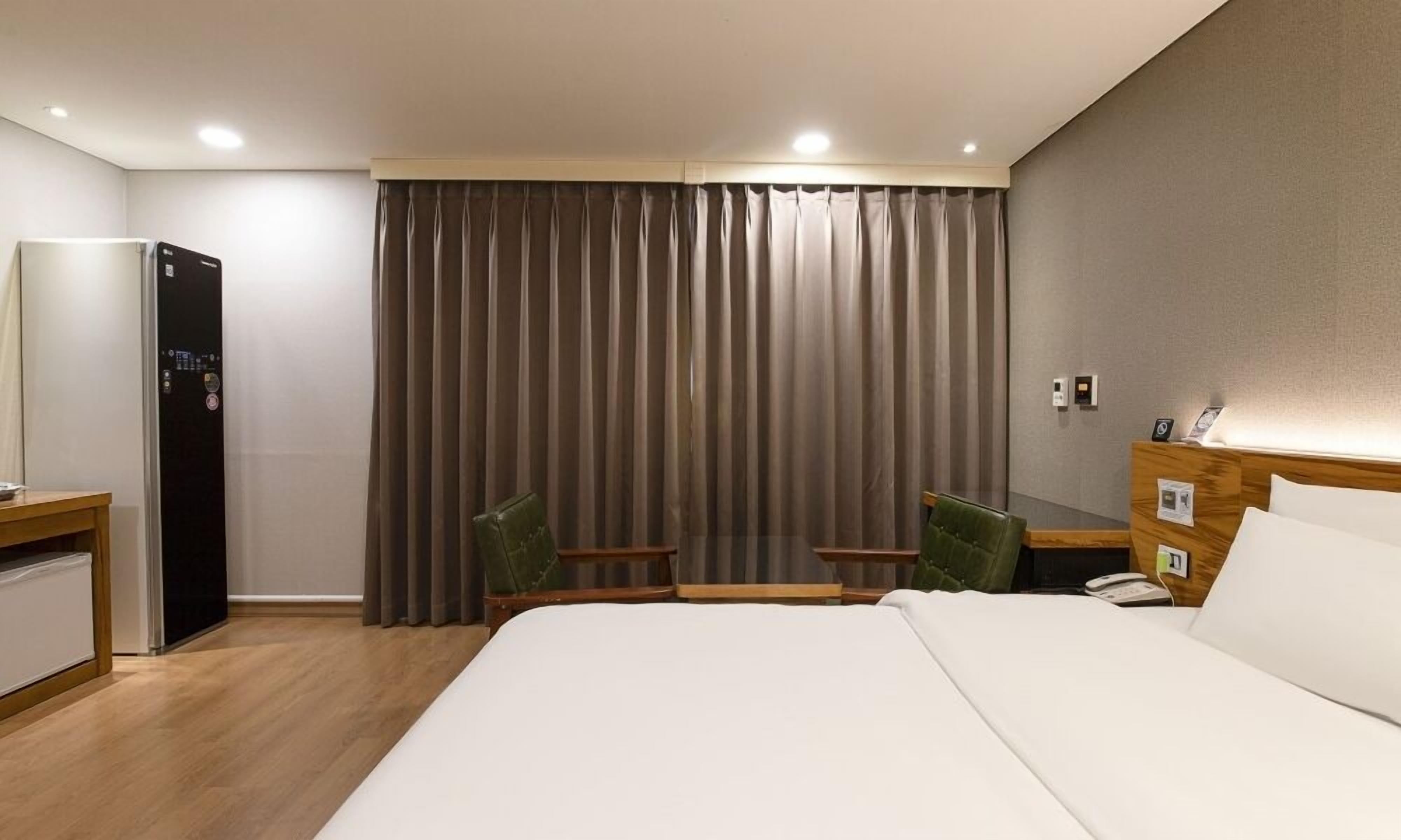 Een hotelkamer met een bed, bureau, stoel, minibar, gordijn en een aan de muur bevestigd telefoon.