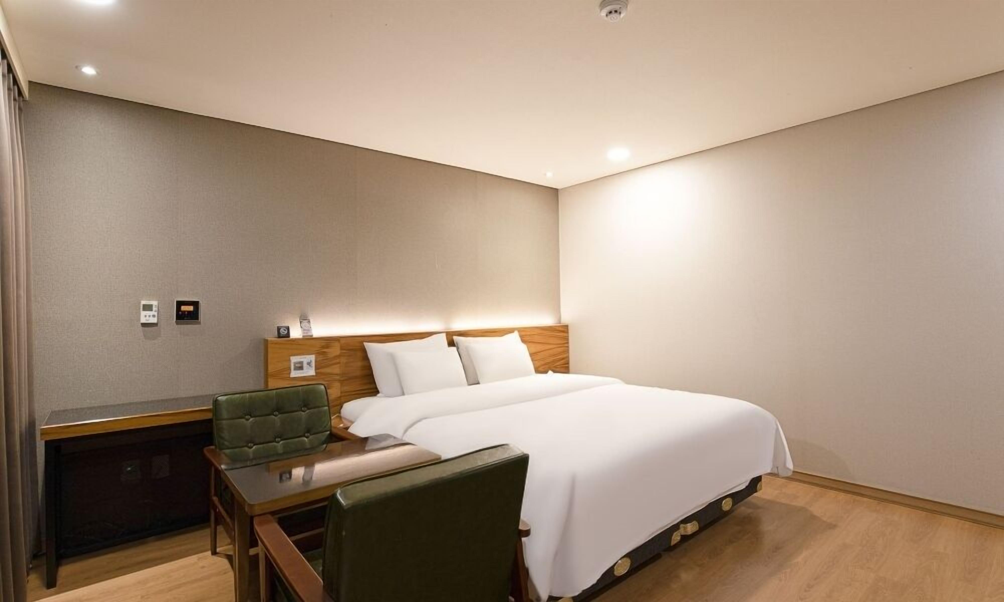 Habitación de hotel con una cama, un escritorio y una silla. Hay dos paneles de control en la pared.