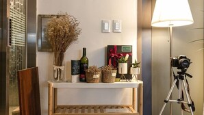 A reception area with a console table displaying a vase of dried flowers, a bottle of wine, a gift box, and potted plants. A camera on a tripod is also visible. - Anyang CNC Hotel (Anyang)