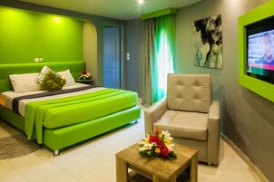 A hotel room with a green bed, a beige armchair, a wooden coffee table with flowers, and a painting on the wall.