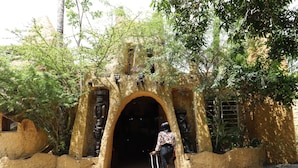 Una persona arrastra una maleta por un arco de entrada con paredes de piedra y un árbol en primer plano.