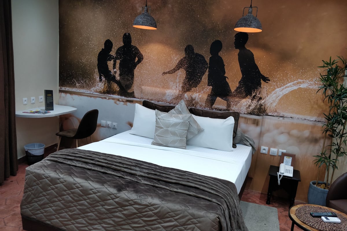 Une chambre d’hôtel avec un lit, un bureau et une chaise. Un mur est décoré d’un tableau représentant des personnes qui se promènent.