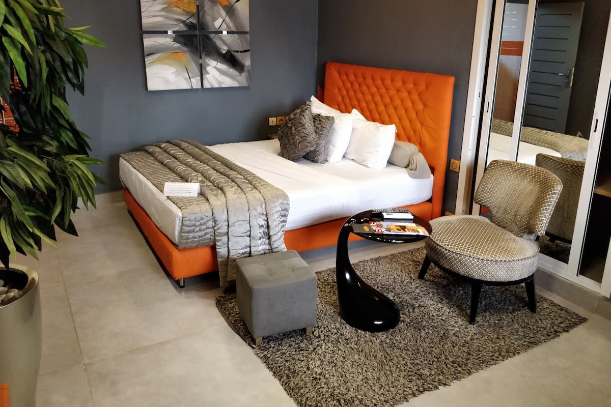 Une chambre moderne avec une tête de lit orange à motifs capitonnés, un banc recouvert d’un tissu gris, une table basse noire et un grand tableau abstrait accroché au mur.