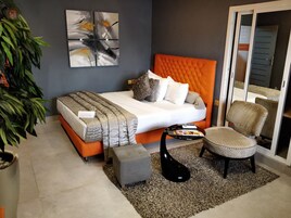 A modern bedroom with a tufted orange headboard, a grey upholstered bench, a black coffee table, and a large abstract painting on the wall.