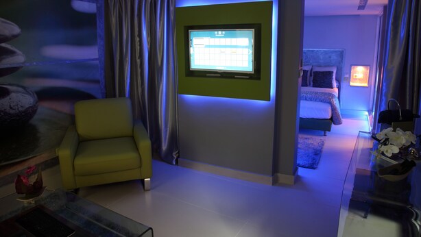 Una habitación de hotel moderna con una televisión de pantalla plana montada en la pared, un sillón amarillo, una cama con cabecero azul y una mesita de noche con un florero.