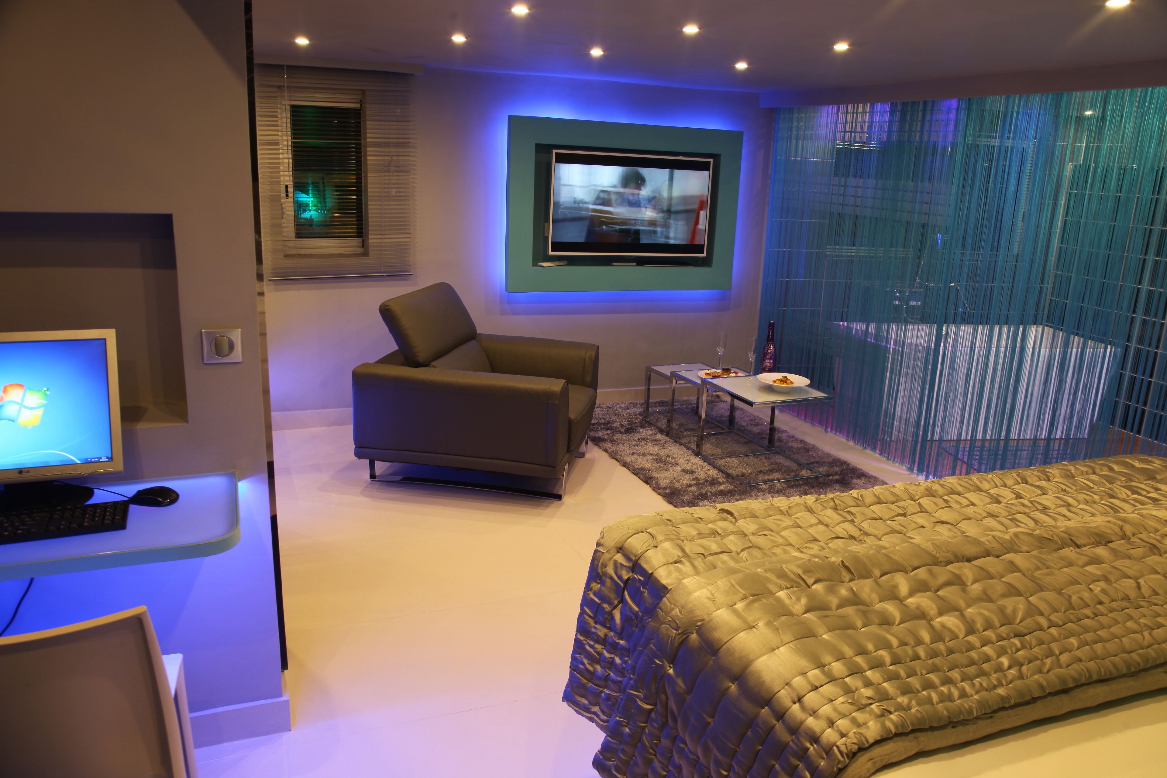 A modern bedroom with a bed, a desk with a computer, a TV, and a seating area with a chair and a small table.