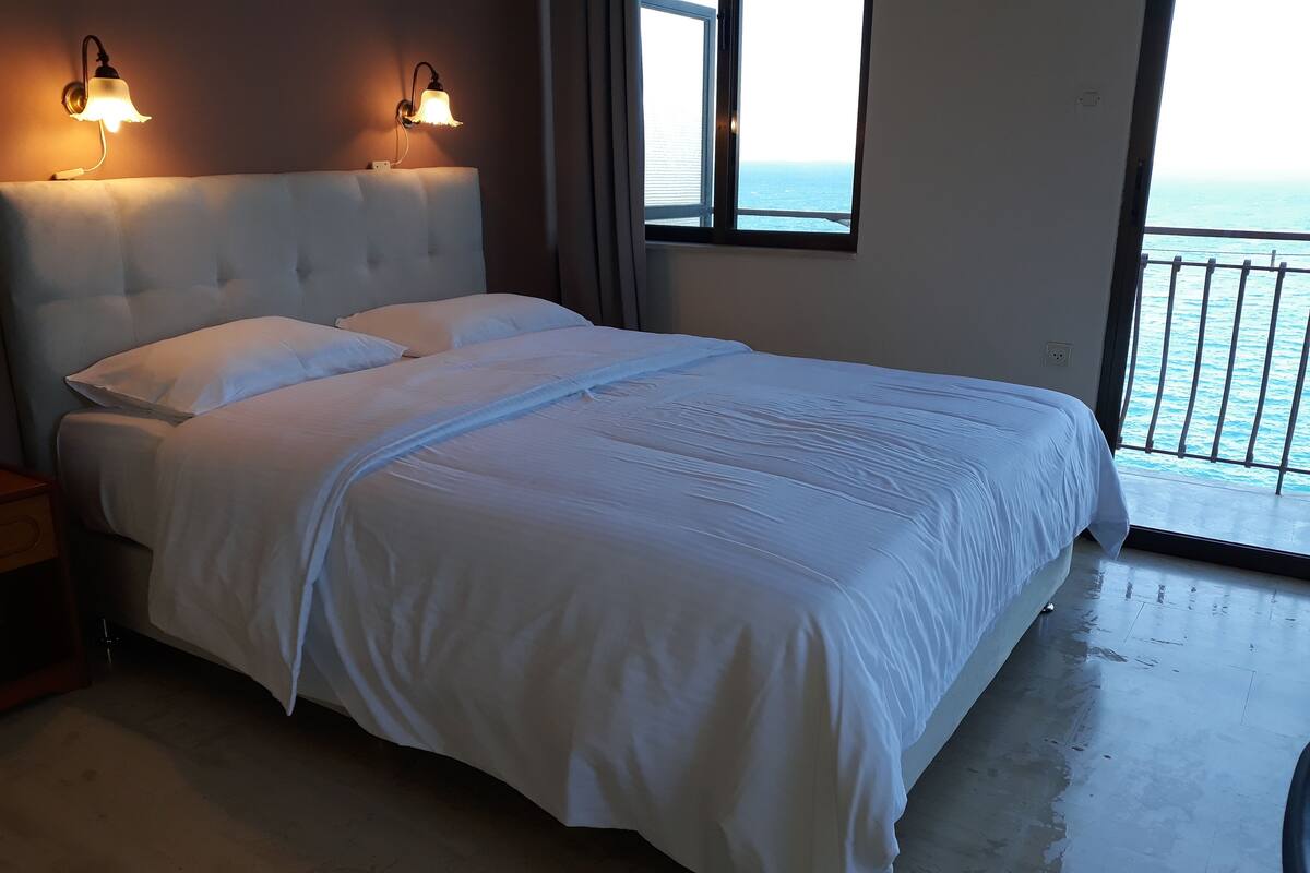 A large bed with white linens, two wall-mounted lamps, and a view of the sea through a window.