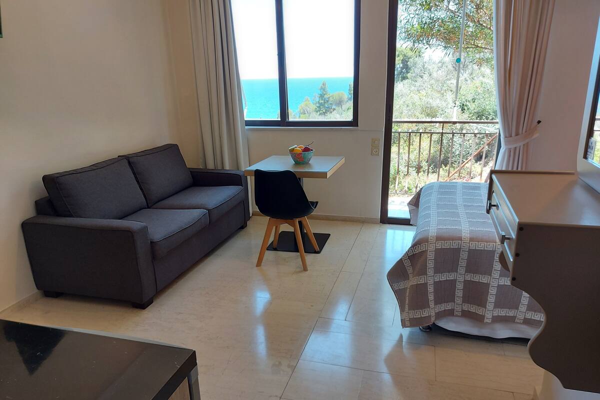 A room with a sofa, a small table with a bowl, a bed with a patterned cover, and a view of the sea through a window.