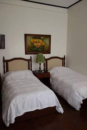 Une chambre avec deux lits simples, une tête de lit en bois, une table de chevet avec une lampe et un tableau représentant des fleurs accroché au mur.