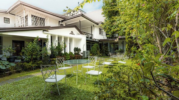 Une cour avec une table en verre et des chaises blanches, entourée d’une végétation luxuriante et d’un bâtiment blanc orné de colonnes.