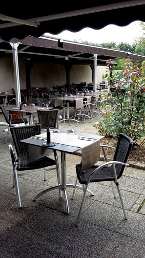 Un patio exterior con mesas y sillas, cubierto por un techo y rodeado de follaje.