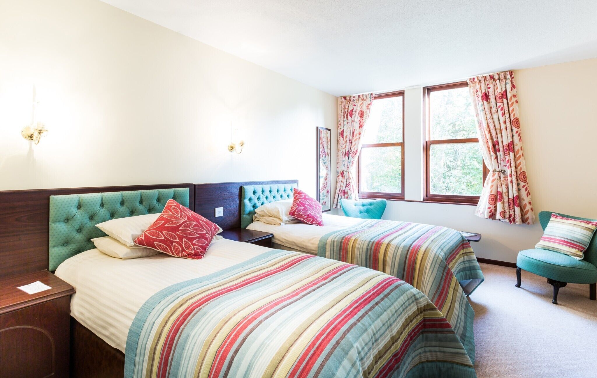 Een tweepersoonskamer met twee bedden, elk voorzien van een gestreept laken, een nachtkastje, een stoel en een raam met bloemen gordijnen.