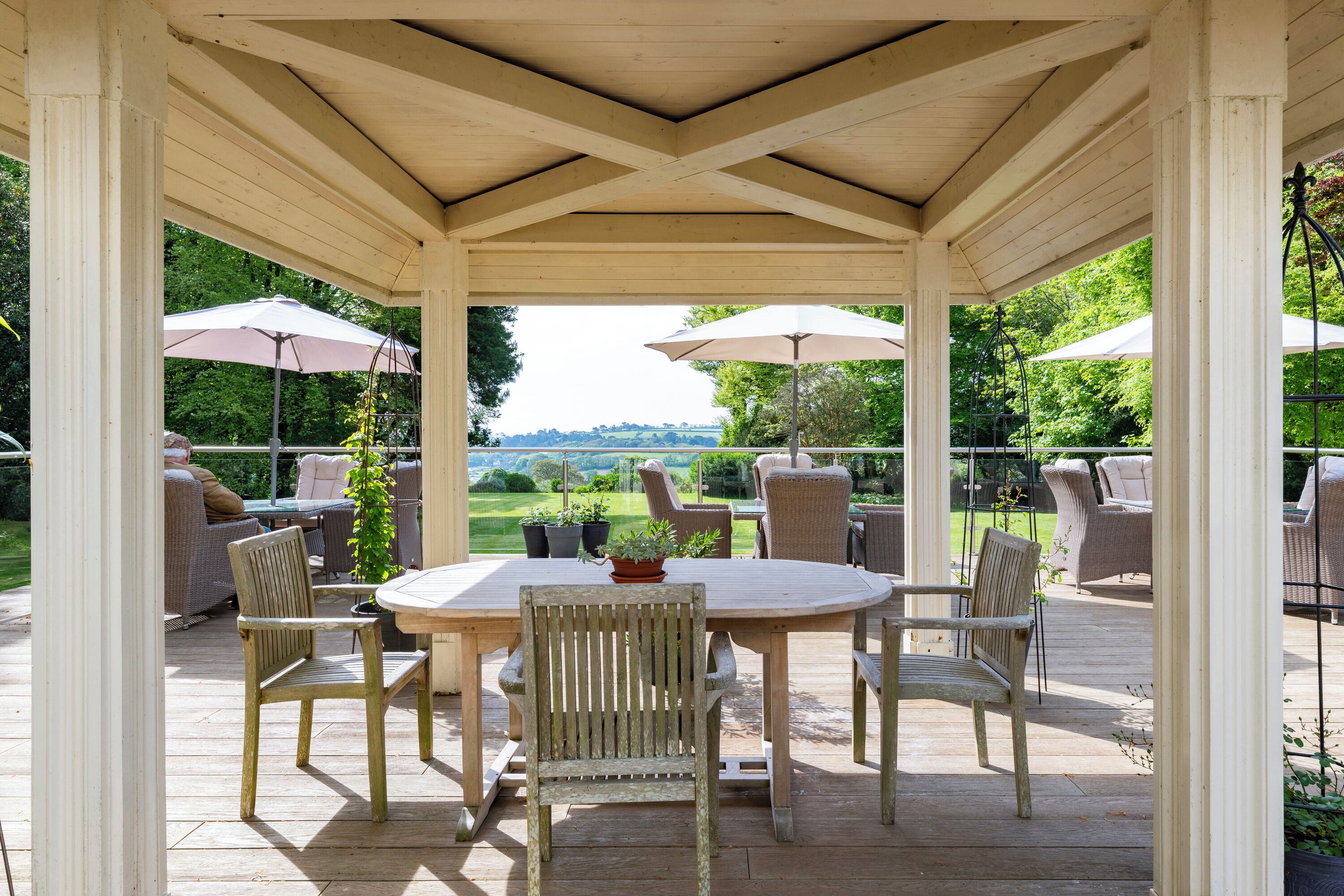 A covered outdoor patio with a round table and chairs, surrounded by potted plants and umbrellas.