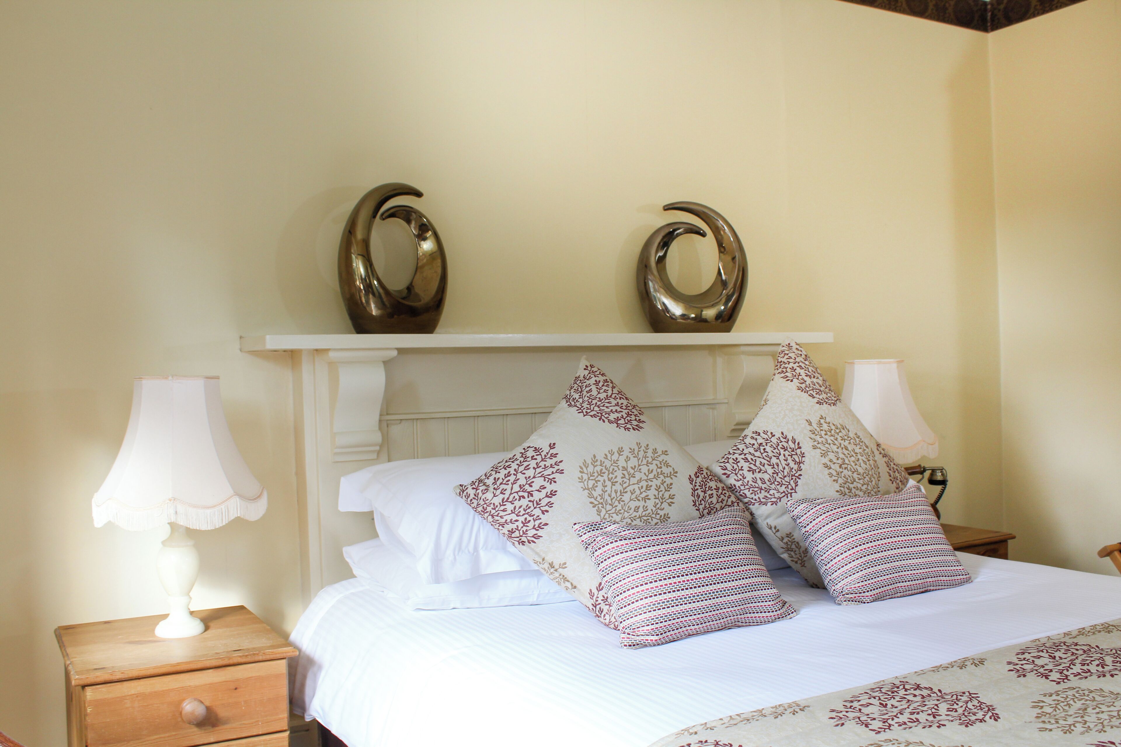Una cama bien tendida con almohadones decorativos, una lámpara de noche y dos espejos fijos a la pared.