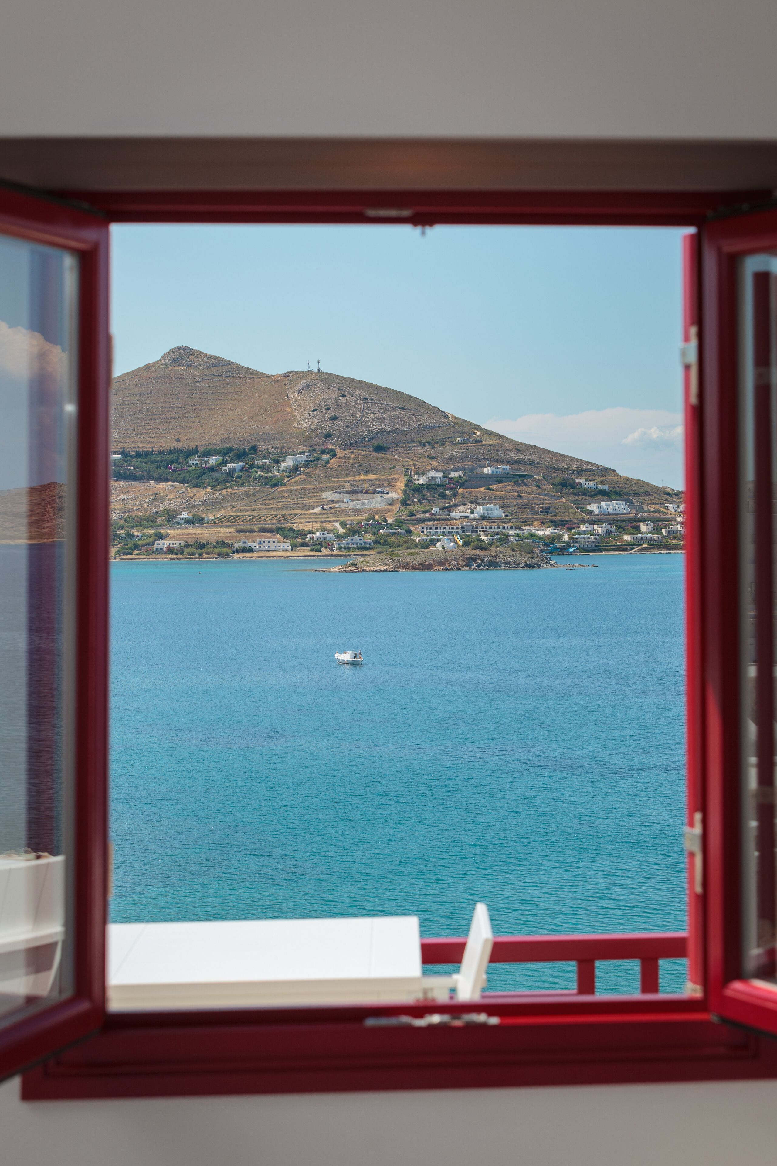 Vista a través de una ventana que muestra un paisaje costero con una montaña, una masa de agua y un barco pequeño.