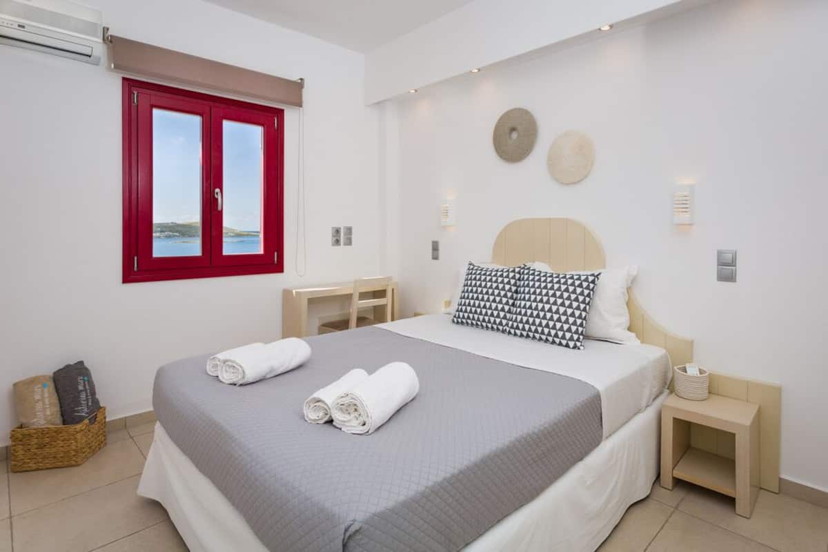 A bedroom with a bed, a desk, a red-framed window, and a view of the sea.