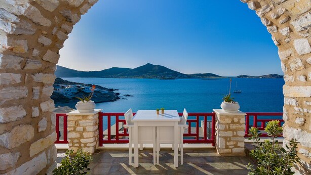 Un arc en pierre mène à une terrasse où se trouvent une table et des chaises blanches, d’où l’on peut admirer une vue imprenable sur la côte, avec ses montagnes et sa mer.