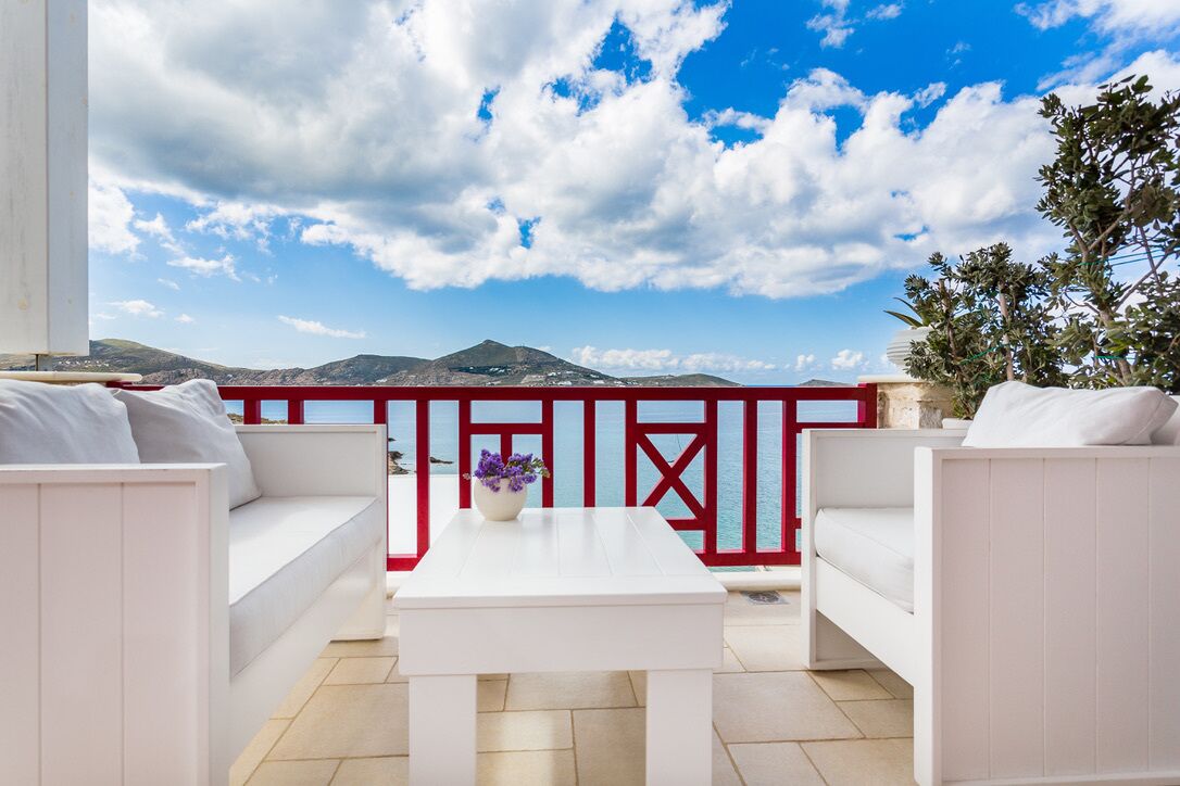 A balcony with white furniture, a red railing, and a view of mountains and the sea.