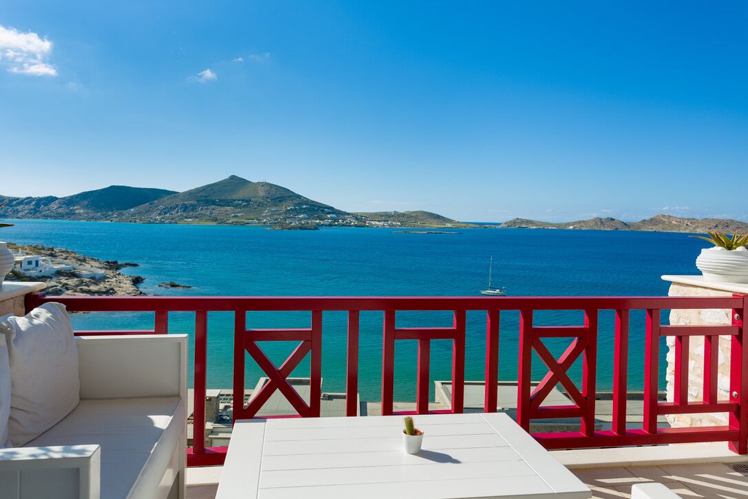 A balcony with a white table and red railing, overlooking a turquoise sea and distant mountains.