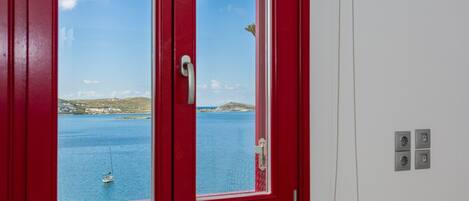 A red-framed window with a view of a calm sea and a sailboat.