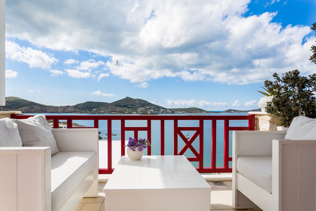 A terrace with white sofas and a table, red railings, and a view of the sea and mountains.