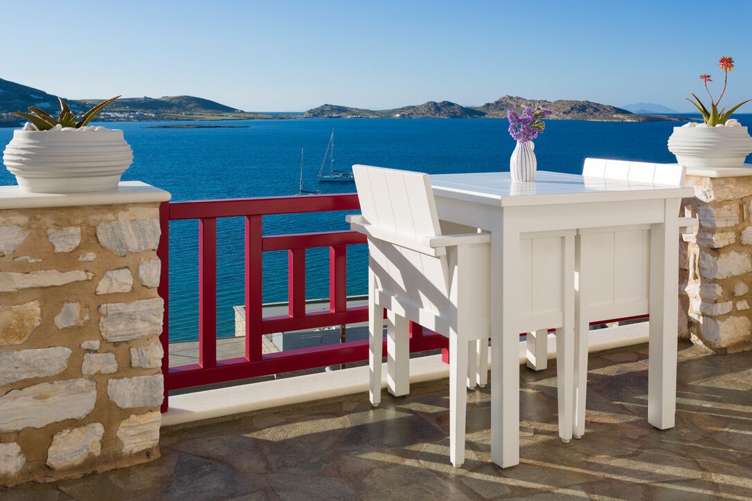 A terrace with a white table and chairs, potted plants, and a red railing overlooking a body of water.