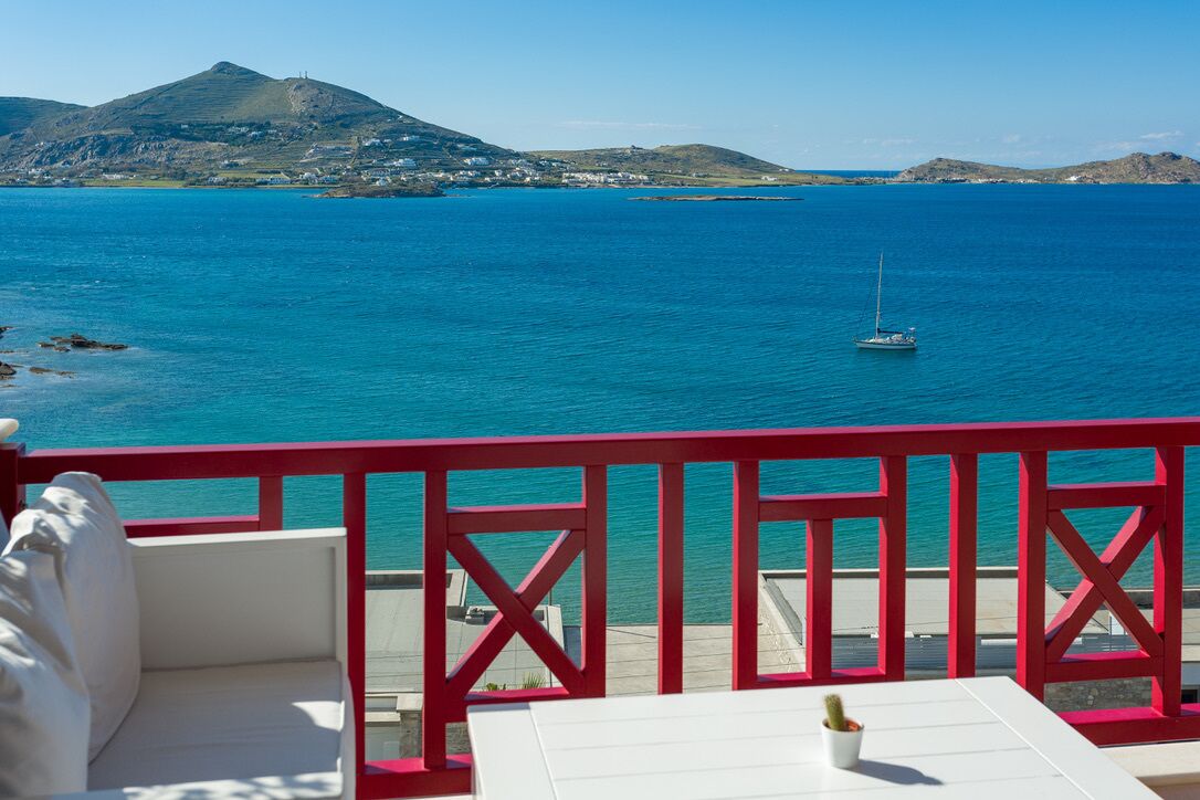 A balcony with a white table and red railing, overlooking a calm sea with a sailboat and distant hills.