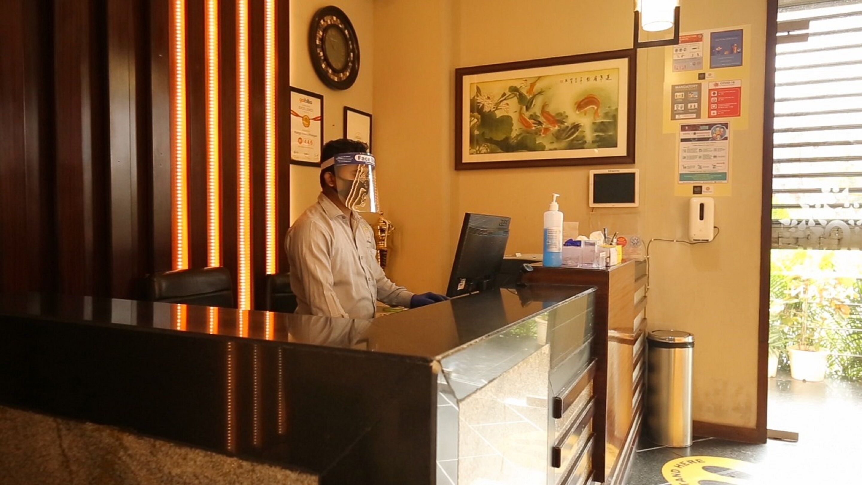 A hotel reception desk with a framed painting, a clock, and informational posters on the wall.