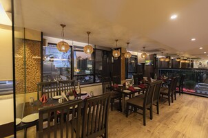 Un restaurante con mesas y sillas de madera, lámparas colgantes y vistas de la ciudad a través de amplias ventanas.