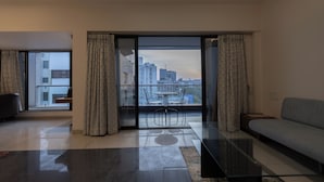 A modern living room with a glass door leading to a balcony, a grey sofa, and a glass coffee table. - Pride Biznotel Alkapuri (Vadodara)