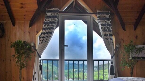 A room with a wooden interior, a balcony with a view of green hills, and two potted plants. - Cottage San Francesco (Nuwara Eliya)