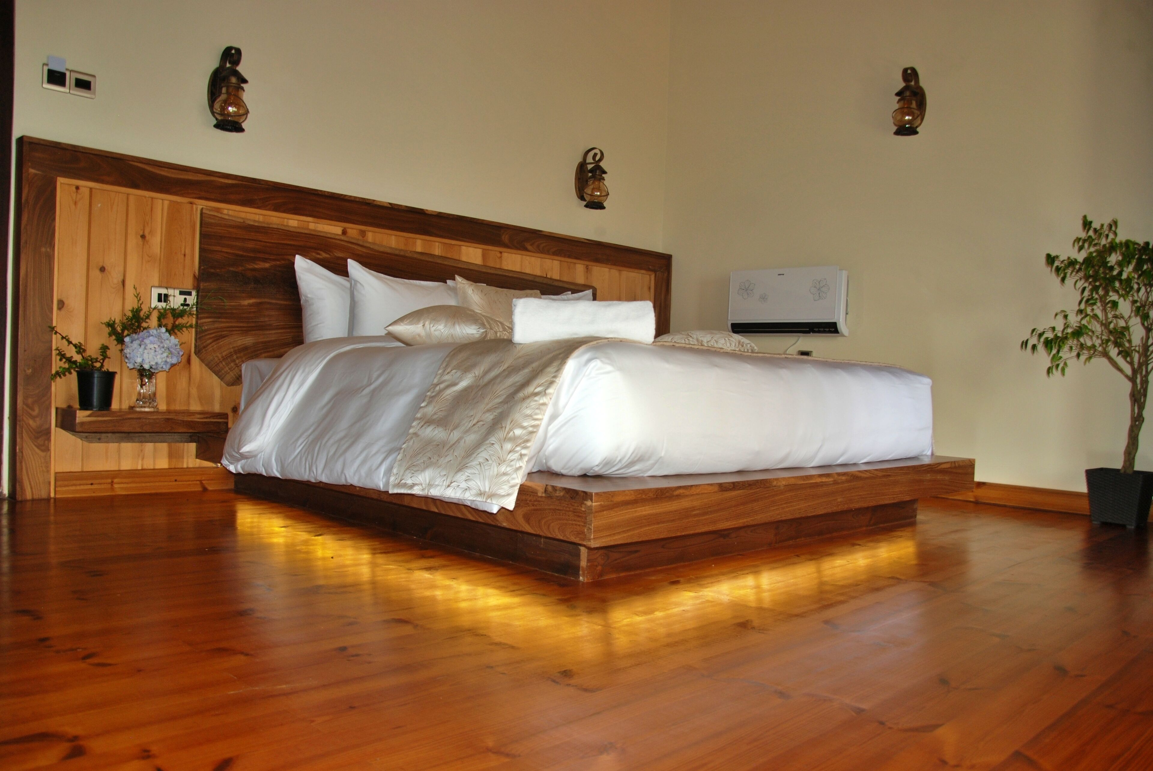 Una cama grande con cabecera de madera, una mesita de noche con un jarrón de flores y una planta en maceta.