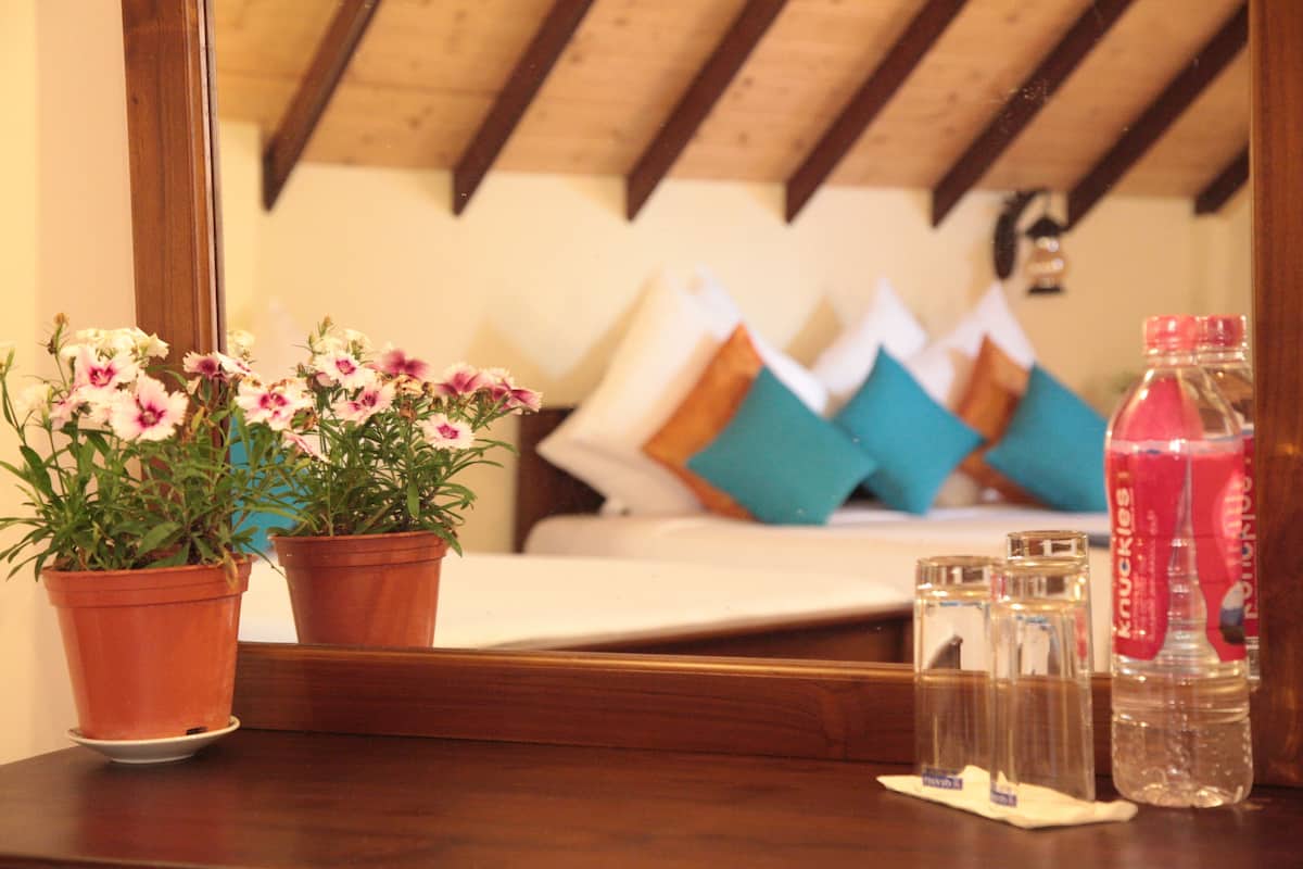 A wooden table with two terracotta pots of flowers, a bottle of water, and a mirror reflecting a bed with pillows.