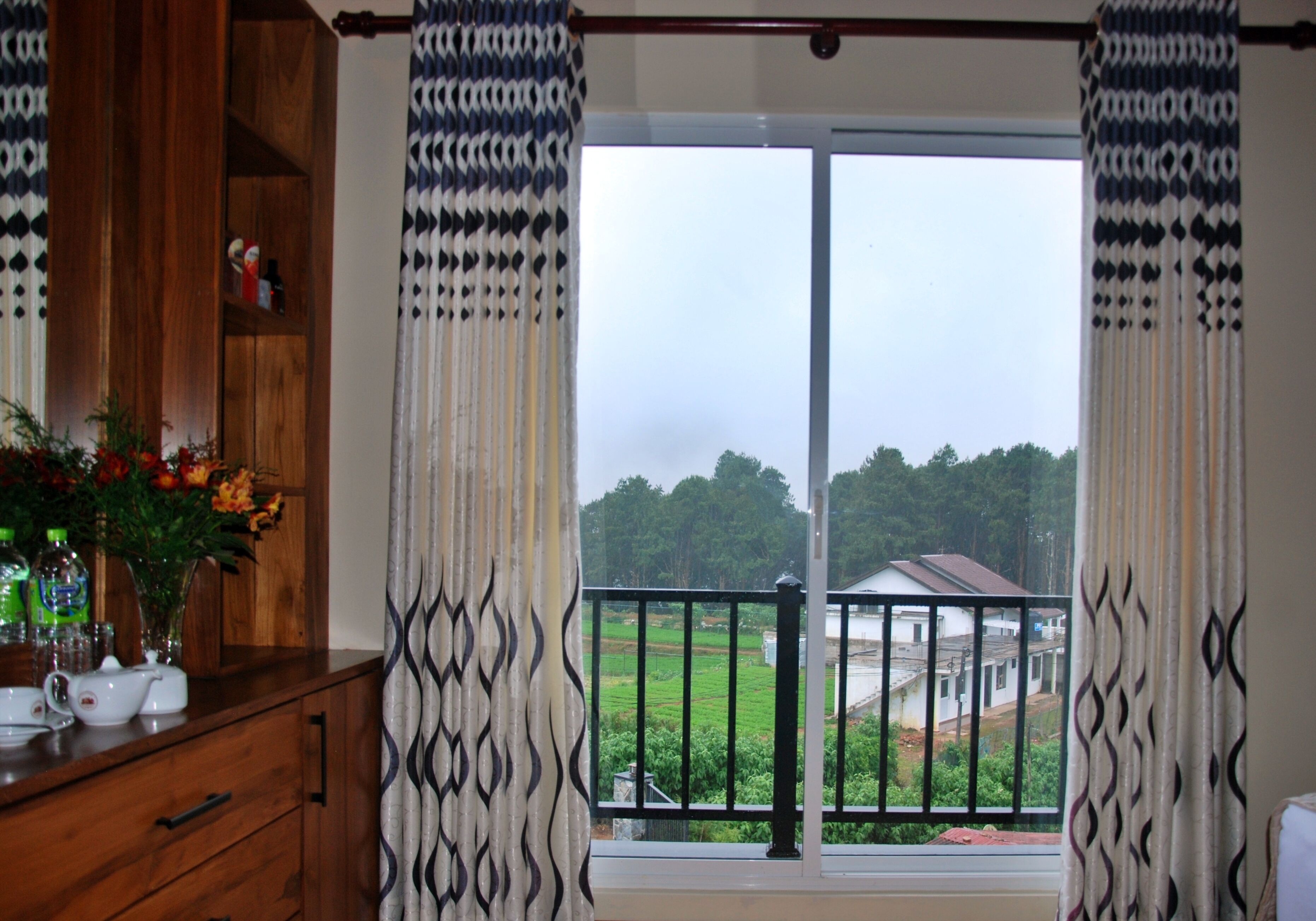 A room with a wooden cabinet, a vase with flowers, and a view of a green landscape through a large window with patterned curtains.