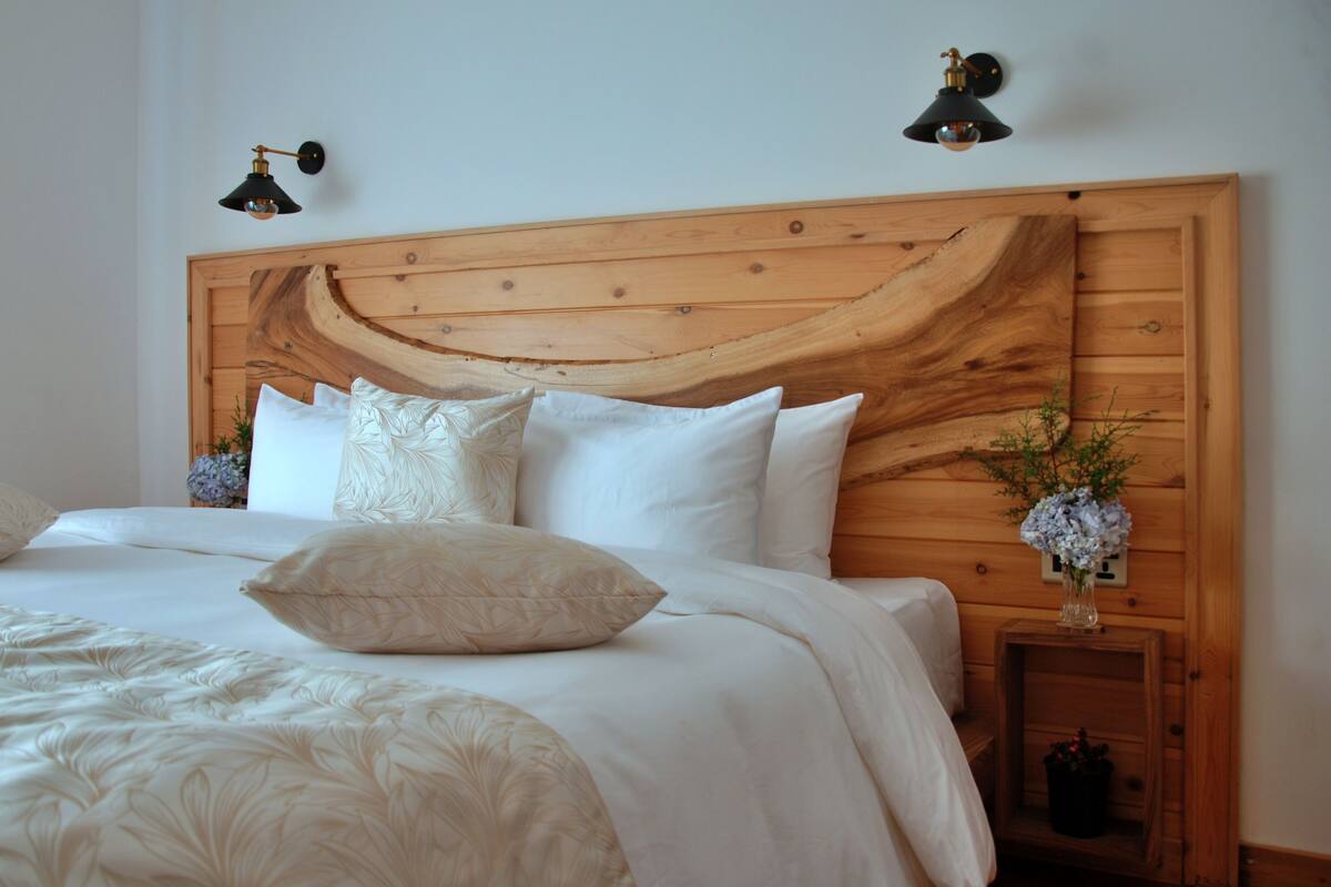 A neatly made bed with white linens and decorative pillows, a wooden headboard, and two wall-mounted lamps.